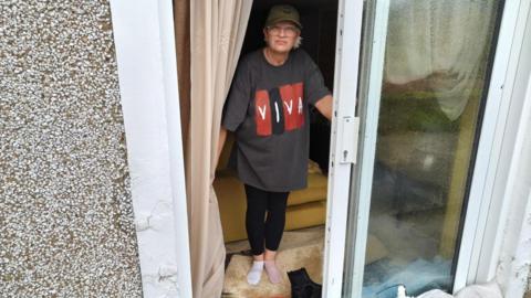 Claire Wheeler, wearing a t-shirt, leggings, a cap and odd socks, stands at the open glass sliding door of her house. There are shoes and clothes piled up by the doorway and a pile of sandbags just outside the door. 