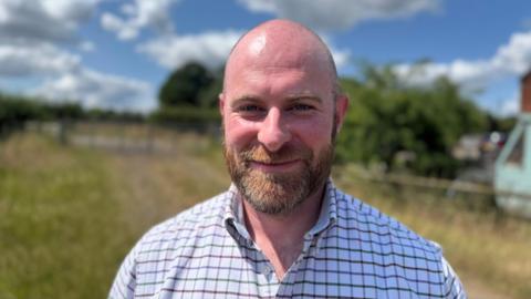 A man with a shaved head and brown beard smiles in a rural field on a sunny day. He is wearing a white shirt with a brown and green checkered pattern.