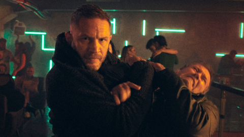 Tom hardy, who has another man in a chokehold. Both men are wearing all black and in the background is a nightclub with people dancing.