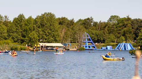 A large lake with trees on the far side and a number of people in boats, with some large inflatible blue slides also on the far side