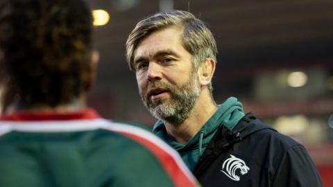 Geoff Parling talking to one of his Leicester Tigers players and he is wearing a green hoodie and black weather proof coat with the Tigers emblem on it.