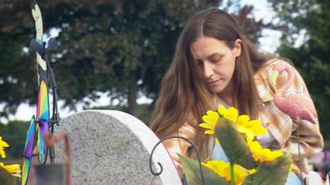 A woman wearing a checked shirt beside a grey gravestone. There are sunflowers next to the gravestone.