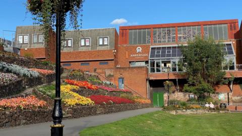 A red brick building, which features a large stained-glass window, is overlooking ornamental flowerbeds and lawns. There is a path leading through the flowerbeds up to the building.