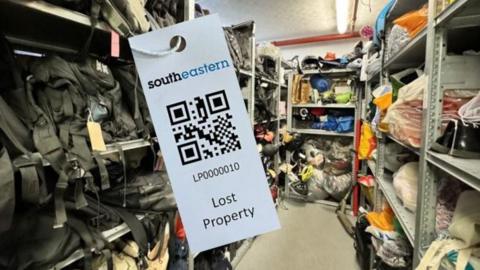 A white 'lost property' tag with a QR code and Southeastern branding hangs off a bag in a room full of collected misplaced belongings. The room is lined of shelves stuffed with shoes, bags, clothing and other items waiting to be returned.