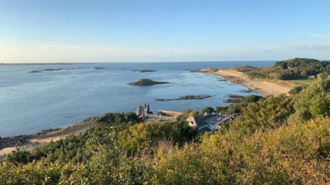 A view over the west coast of Herm, with a beach, buildings, trees, the sea and islets in view on a sunny day.