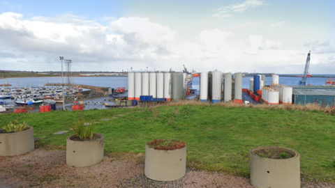 Peterhead Harbour in front of blue skies and cloud. In the front of the picture there's green grass and planters. The harbour has a number of boats and structures. 
