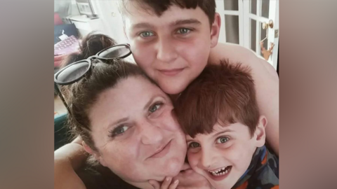 A woman and two young boys huddle together and smile into the camera. The woman has dark hair pulled back from her face and dark framed glasses perched on her head. The two boys have brown hair.