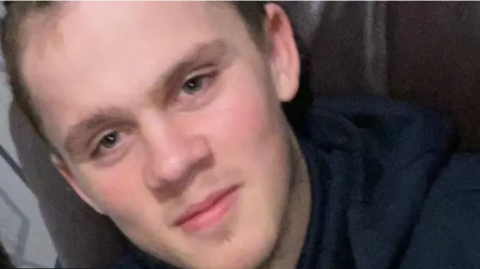 A close-up of a man's face. He is looking at the camera and has brown hair and is wearing a black top. 