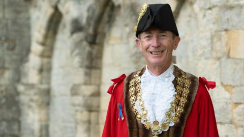 A man in a town mayoral cloak and hat is standing in front of a stone wall. He is smiling at the camera.