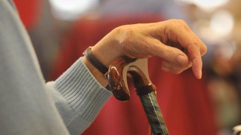 A stock image showing a person's hand on a walking stick. They are wearing a pale blue jumper and a watch.