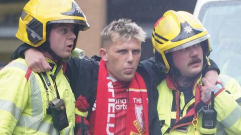 Jack Trotter, who has short blonde hair and is wearing a Liverpool FC shirt and scarf, has his arms around two firefighters who are helping him to walk. They are wearing high-viz jackets and yellow helmets