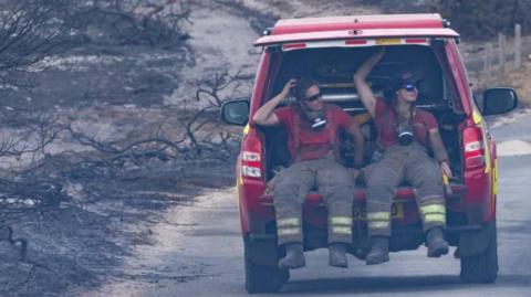 Two fire officers, one female, are in the back of a red fire vehicle with the boot door open. They are wearing fire safety clothing and around their necks are breathing masks. Both wear sunglasses. Around them are the remnants of burnt trees and grassland. The red vehicle appears to be driving on a road away from the camera.