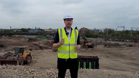 A still from a video shows a man in a white hard hat and hi-viz vest talking to the camera against a backdrop of rubble  with two yellow diggers. In the background, overhead electric power lines run along a railway line. 