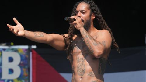 Bobby Vylan without his shirt on stage at Glastonbury 2025. He has a blackbeard and long dreadlocks