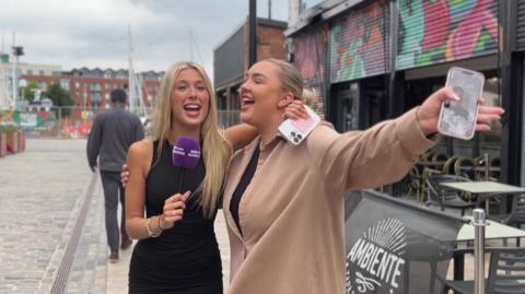 Two women, with their arms around each other, sing in the street. One holds a purple mic