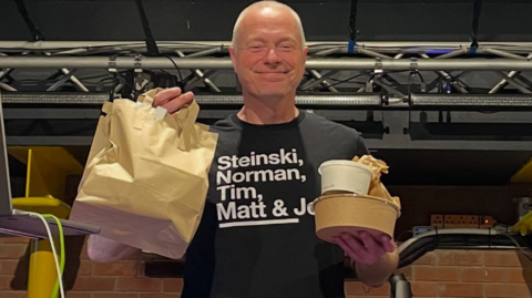 Aldo Vanucci is smiling, wearing a black T-shirt that reads 'Steinski, Norman, Tim, Matt' and holding a brown paper bag and bowls of takeaway food.