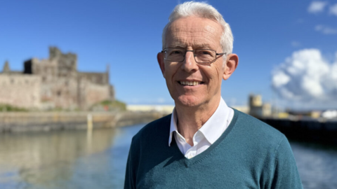 Graham McAll has short white hair and wears glasses. He's wearing a blue jumper over a white shirt. He is smiling and standing in front of a harbour, with castle ruins to the left and it is a sunny day. 