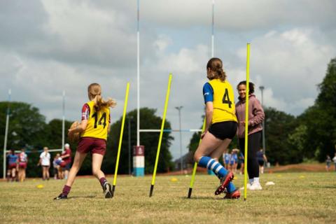 Two girls in bibs run between poles, with one carrying a rugby ball. A woman in a tracksuit watches over them.