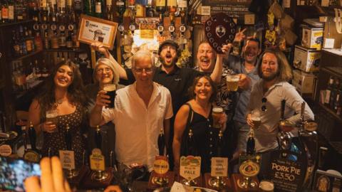 Staff behind the bar at The Blue Bell in York - a mixed gender group of people stand in a traditional pub, with low lighting.