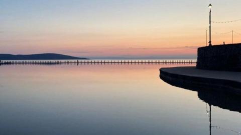 Marine lake in Weston-super-Mare during sunset. There is a concrete walkway to the right, with a large stone wall, which features a lampost on it. A mountain/hilly figure can be seen in the distance.