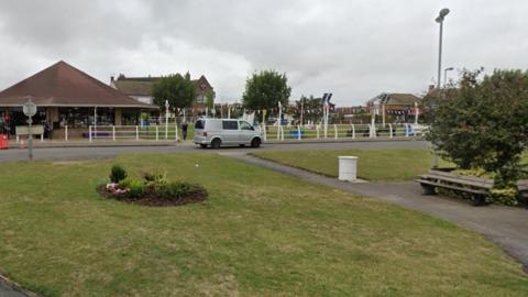 A Google view of the area around The Green in Chapel St Leonards. There are flowers, benches and a shop in the background, and a van is driving past.