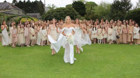 A bride, with blonde hair, a white dress with an off the shoulder detail carrying a bouquet, smiles as two young bridesmaids help her with her train and veil as she walks towards the camera. Behind them is a large group of women, including young girls in the front row, wearing a mixture of champagne, cream, white and tan dresses. The crowd, including the bride and the two bridesmaid, are standing in the middle of a large garden, with tall trees and a leafy tunnel to the left.