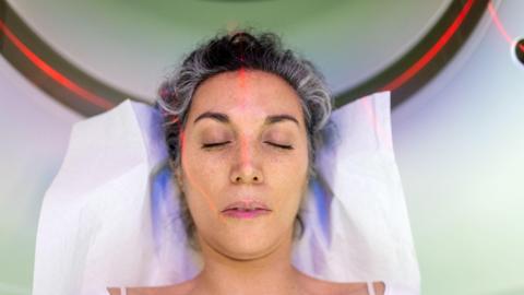 I woman with her head on a white pillow, she is having her head scanned by a piece of medical equipment with red lazers