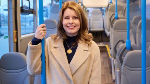 North East Mayor Kim McGuinness. She has shoulder length brown hair, a beige coat, black top and is wearing a gold necklace and two rings. She is holding on to a metal pole on the bus and is smiling. There are empty, grey seats on the bus.