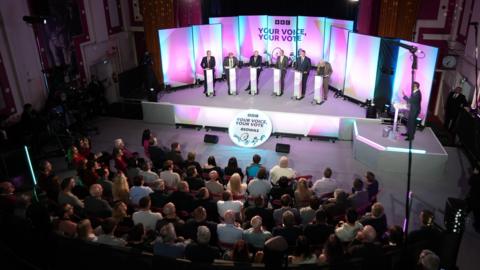 The Caerphilly by-election candidates stand on stage with individual podiums, they are all men wearing suits. The audience is visible, seated facing the stage.