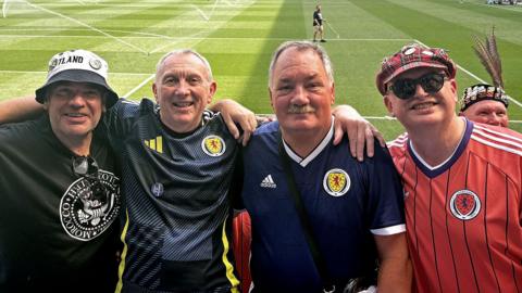 Four men with their arms around each others' shoulders inside a football stadium, with the pitch and other stands visible behind them. The two men in the middle are wearing blue Scotland tops. The man on the left is wearing a black t-shirt and a bucket hat, and the man on the right has a salmon pink Scotland top and a tartan cap and sunglasses.