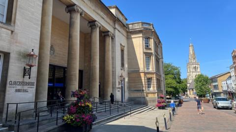 The front entrance to the Gloucestershire Shire Hall has four pillars in front of it and some flowers on the side. There are people walking past it on the street in front.