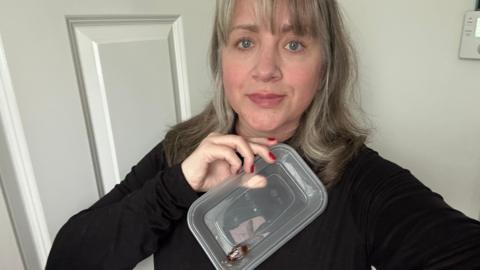 Amy with mousy brown hair and wearing a black jumper is holding up a plastic box with a dead cockroach inside