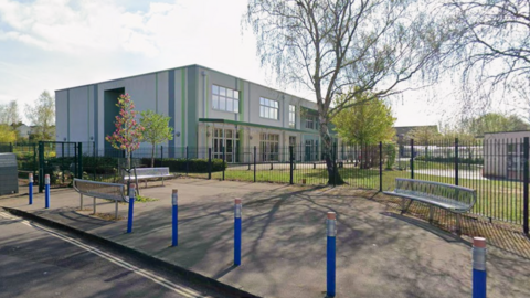 A screenshot from Google street view showing the exterior of May Park Primary School. It is a grey-white rectangular building with green and blue stripes and large windows. There is a small green space with trees growing in front of the building. Outside the metal fencing, there are blue bollards shaped to look like pencils spaced out evenly on the pavement.