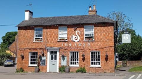 The building of a pub. It has The Swan written on it and an image of a swan too. It has a off-white front door and a car parked to the left of the building
