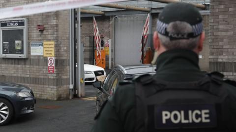 A police officer stands with his back to the camera and is stood near a grey, unmarked police car. There is a police cordon closing off the station and a white Audi car is visible in the distance in the station, flanked by two large grey brick blast walls.