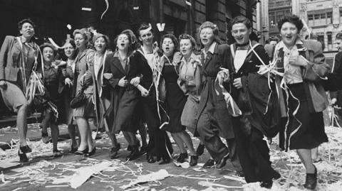 Men and women are lined up singing and dancing and covered in streamers as celebratations are held on VE day in this black and white archive photograph. The women are wearing 1940s skirts, jackets and blouses and two of the men are in uniform. There are reams of streamers underfoot.
