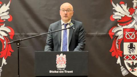 Stoke-on-Trent City Council chief executive Jon Rouse. He is stood at a black podium, against a black back drop - both of which bear the council's logo which is a red and white crest. Mr Rouse is wearing glasses, a black suit with a white shirt and a patterned blue tie.