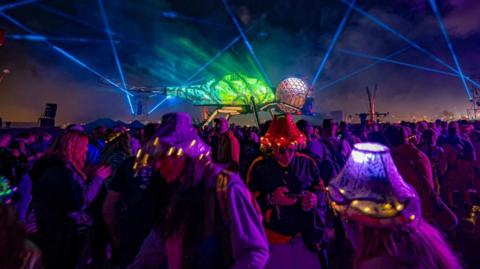 A biomechanical dragonfly lit up in blue and green with thousands of people dancing underneath.