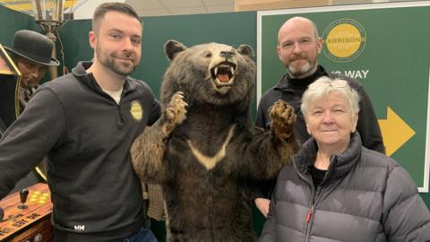 A man with short dark hair and stubble is wearing a black jumper. He is standing next to a 6ft taxidermy grizzly bear in an antiques centre. Also stood next to the bear is a man with a bald head and short beard in a black jumper and a woman with white hair and glasses on the top of her head in a grey puffer coat.