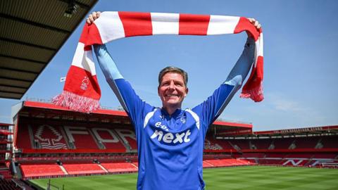 Oliver Glasner faces into the sunshine holding a red and white bar scarf, with the City Ground pitch and stands in the background