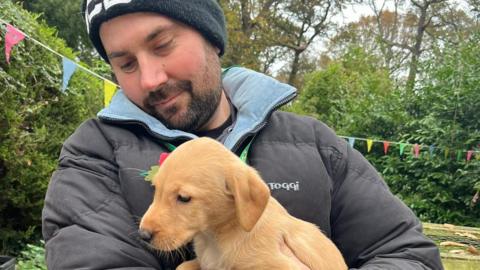Robin Elcock is dressed in a black puffer jacket and a black beanie hat. He is looking down at a puppy he is holding, smiling.