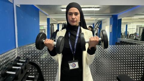 Nadia Ali - a woman wearing a white blazer and black top - is holding up two black weights in a gym. There are mirrors behind her.