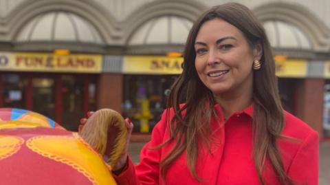 Serena Silcock has long brown hair, she is wearing a red jacket and gold earing and is stood outside Southport Funland