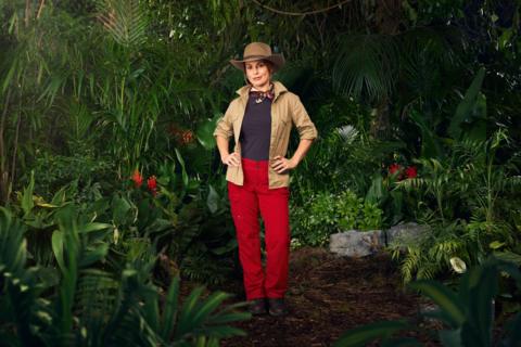 Coleen Rooney wears red cargo pants, a blue t shirt and khaki shirt, as well as a hat as she stands in a jungle