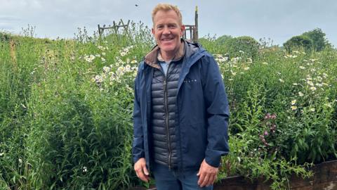 Adam Henson, wearing all navy blue - blue jeans and a blue waterproof - stands in front of a tall bed of wildflowers