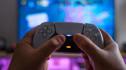 A white PlayStation 5 controller being held up by someone's hands against a blurred TV screen behind it