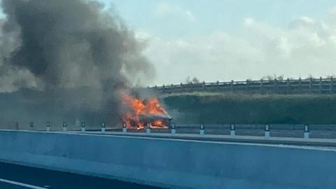 The car engulfed with flames resulting in thick black smoke rising. The car is parked on the side of the road. The photo is taken from the opposite carriageway.