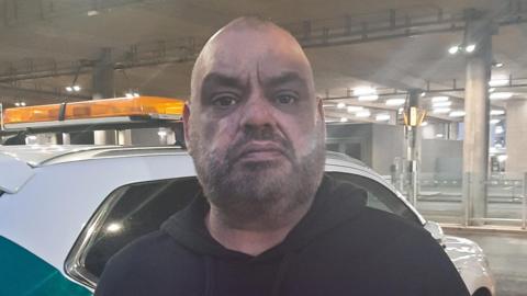 Man in black hoodie next to a parked car in front of an airport 