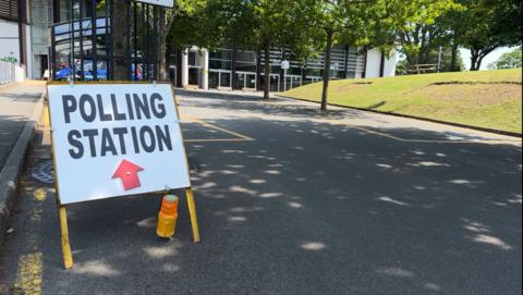 A sign which says polling station with a red arrow pointing towards Beau Sejour, a leisure centre with trees outside and some greenery on the right. 