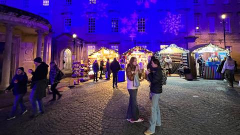 People gathered outside chalets on an evening at Bath Christmas Market, with Christmas lights on the chalets and the buildings behind illuminated in blue